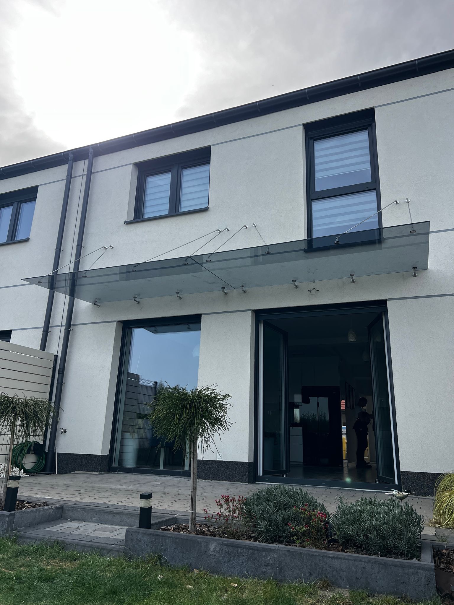 Szklany daszek nad wejściem do nowoczesnego domu z otwartymi drzwiami balkonowymi. Minimalistyczny design, szklana konstrukcja wsparta na metalowych elementach.