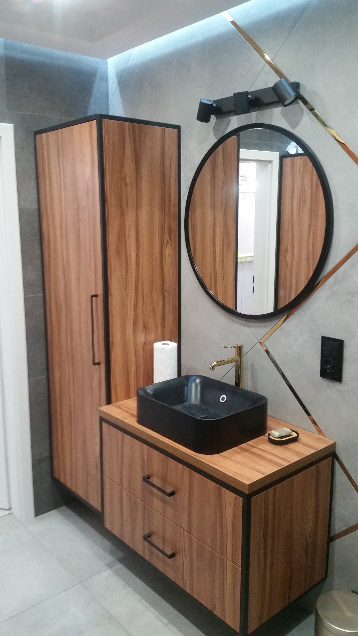Nowoczesna aranżacja łazienki z drewnianą szafką pod umywalkę, czarną, prostokątną umywalką nablatową, okrągłym lustrem z czarną ramą i złotymi akcentami na szarej ścianie.
