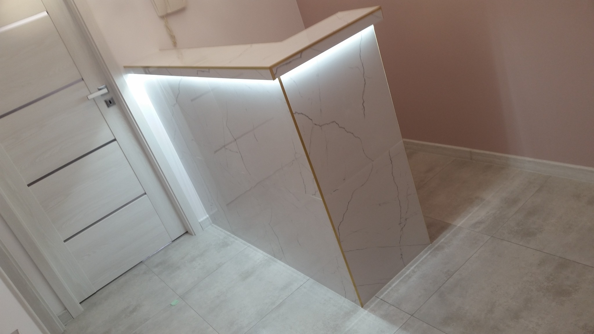 Wykończony fragment wnętrza z zabudową przypominającą barek lub wyspę kuchenną, wykończoną płytami imitującymi marmur z podświetleniem LED, widoczna biała ściana i drzwi z poziomymi frezami.