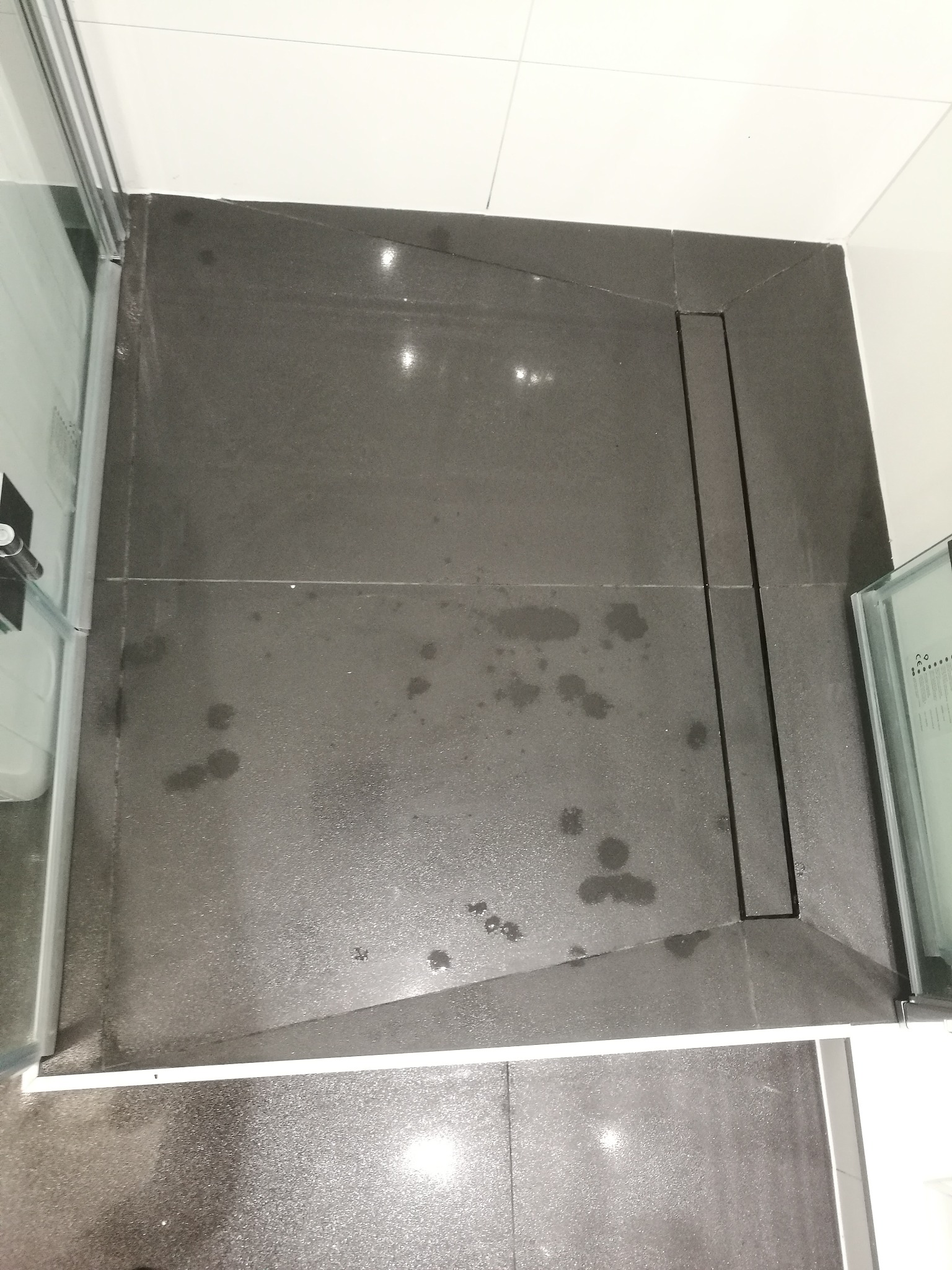 Szare płytki podłogowe w kabinie prysznicowej z odpływem liniowym, widoczne ślady po wodzie, białe fugi i fragment szklanej ścianki kabiny.