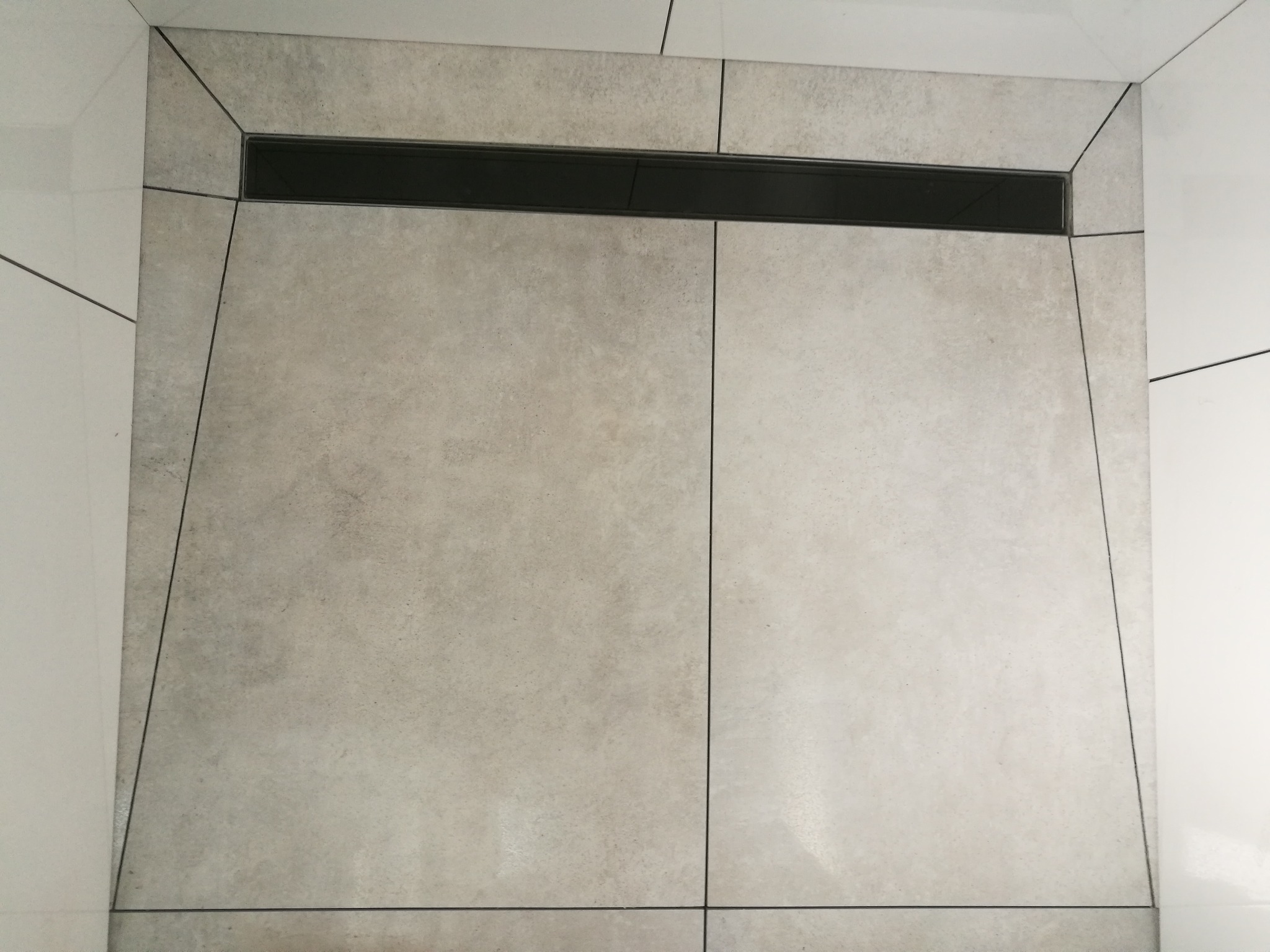 Nowoczesna łazienka z odpływem liniowym w podłodze wyłożonej szarymi, matowymi płytkami o dużym formacie, kontrastującymi z białymi ścianami.