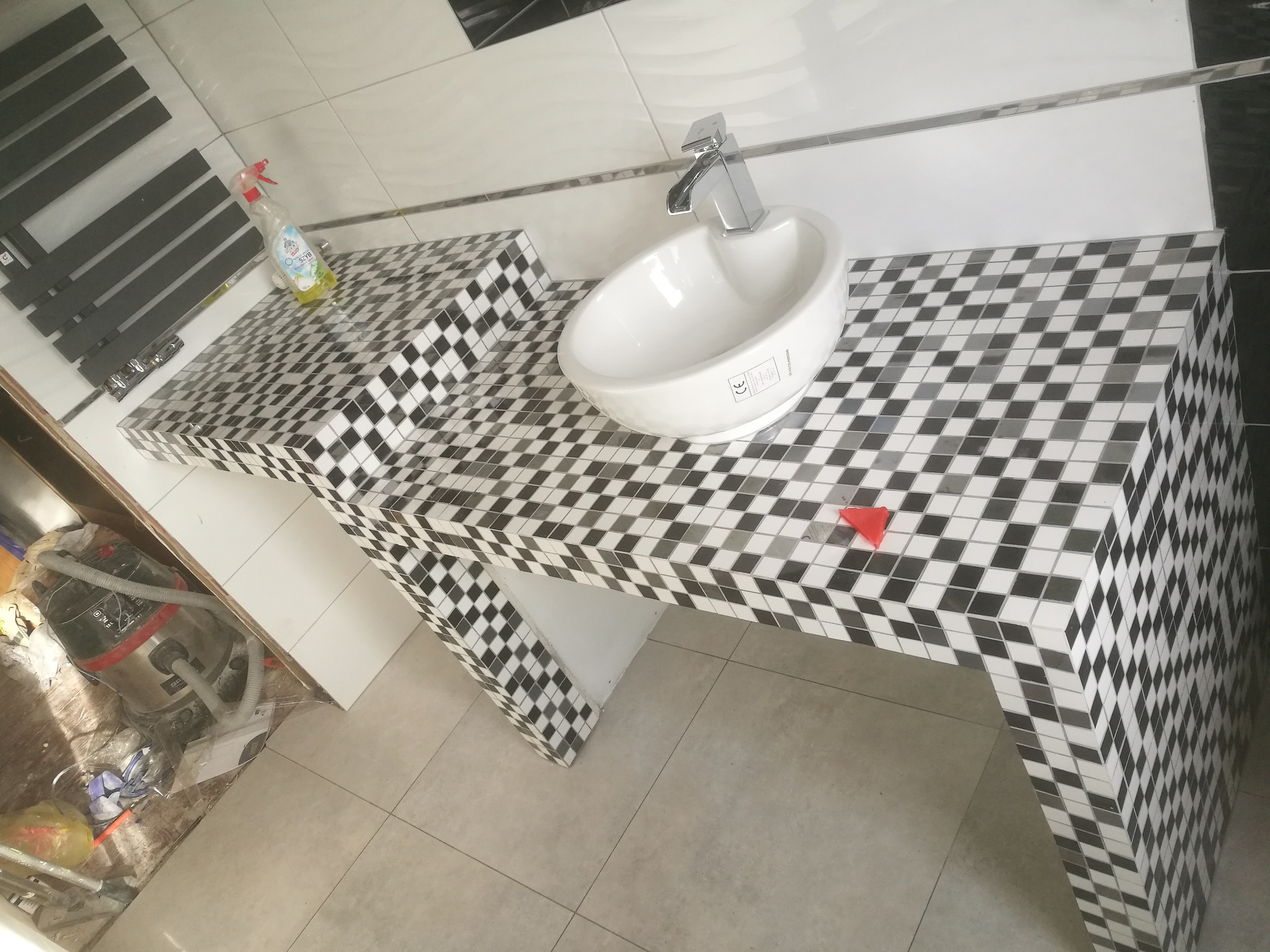 Umywalka nablatowa w łazience wykończonej mozaiką czarno-białą, widoczny fragment podłogi z płytek oraz grzejnik drabinkowy i butelka ze środkiem czyszczącym.