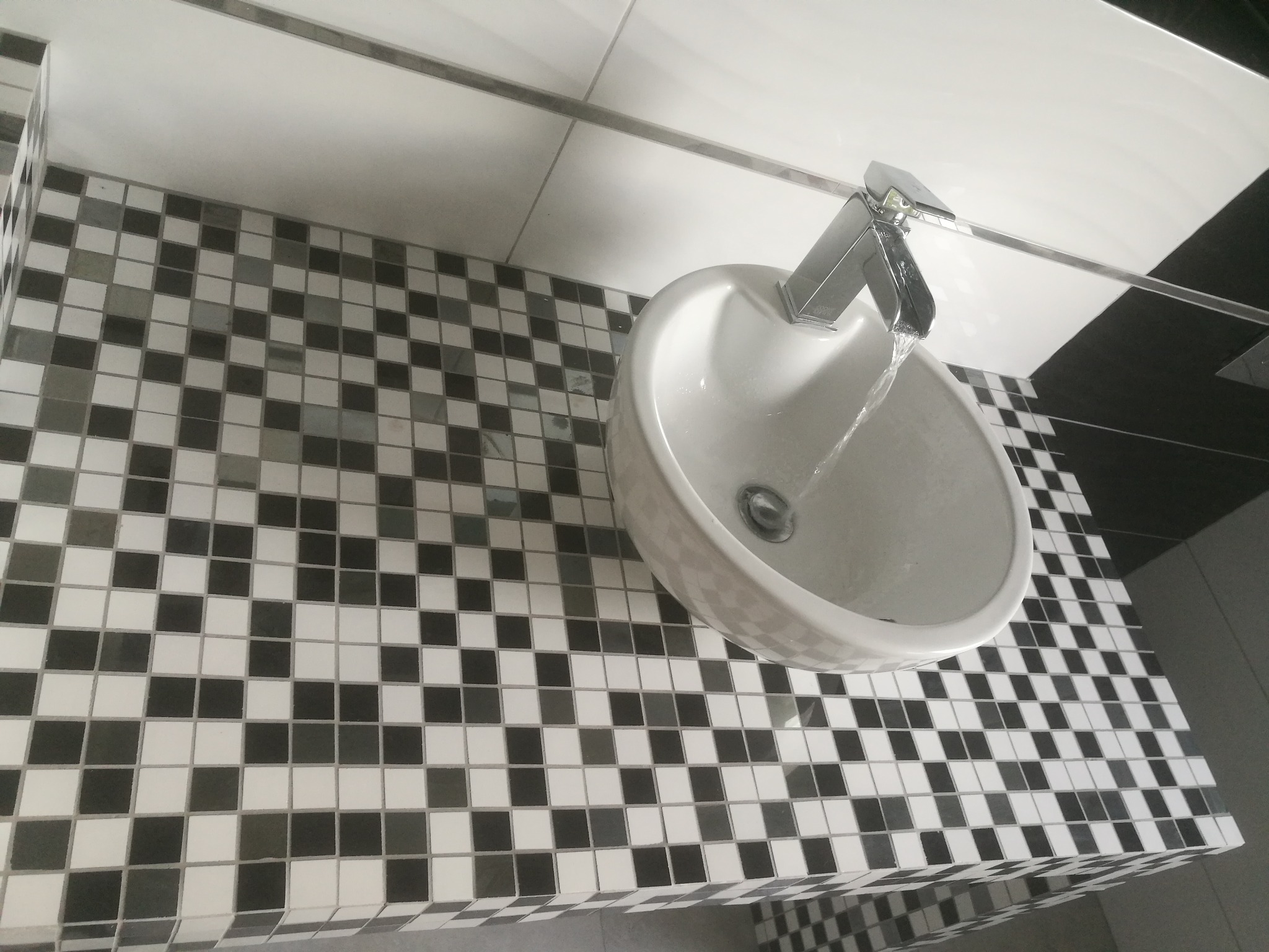 Biała, okrągła umywalka z chromowaną baterią, z której płynie woda, umieszczona na blacie wyłożonym mozaiką z czarno-białych kwadratów. W tle widoczna biała ściana z czarnym paskiem.