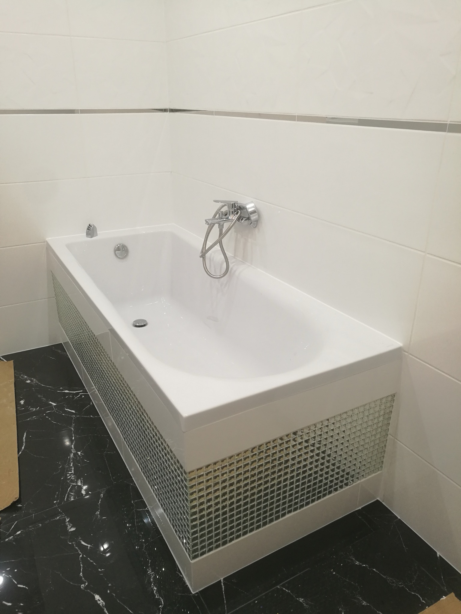 Nowoczesna, biała wanna z mozaikową obudową i chromowaną armaturą, osadzona w łazience z białymi płytkami ściennymi i czarną marmurową podłogą.