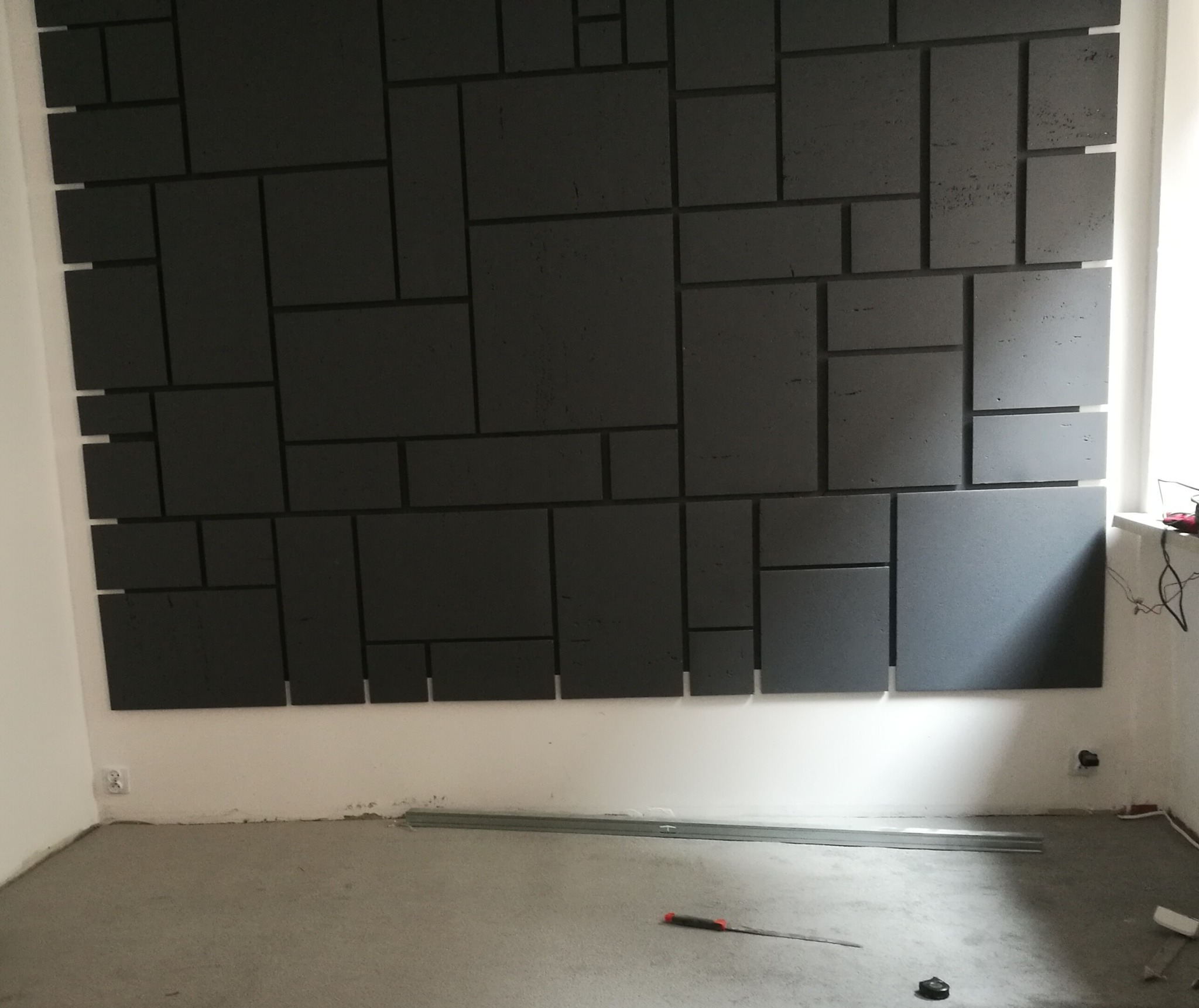 Ściana z zamontowanymi, ciemnoszarymi panelami o zróżnicowanych rozmiarach, tworzących geometryczny wzór, widoczne narzędzia na podłodze, w tle okno.