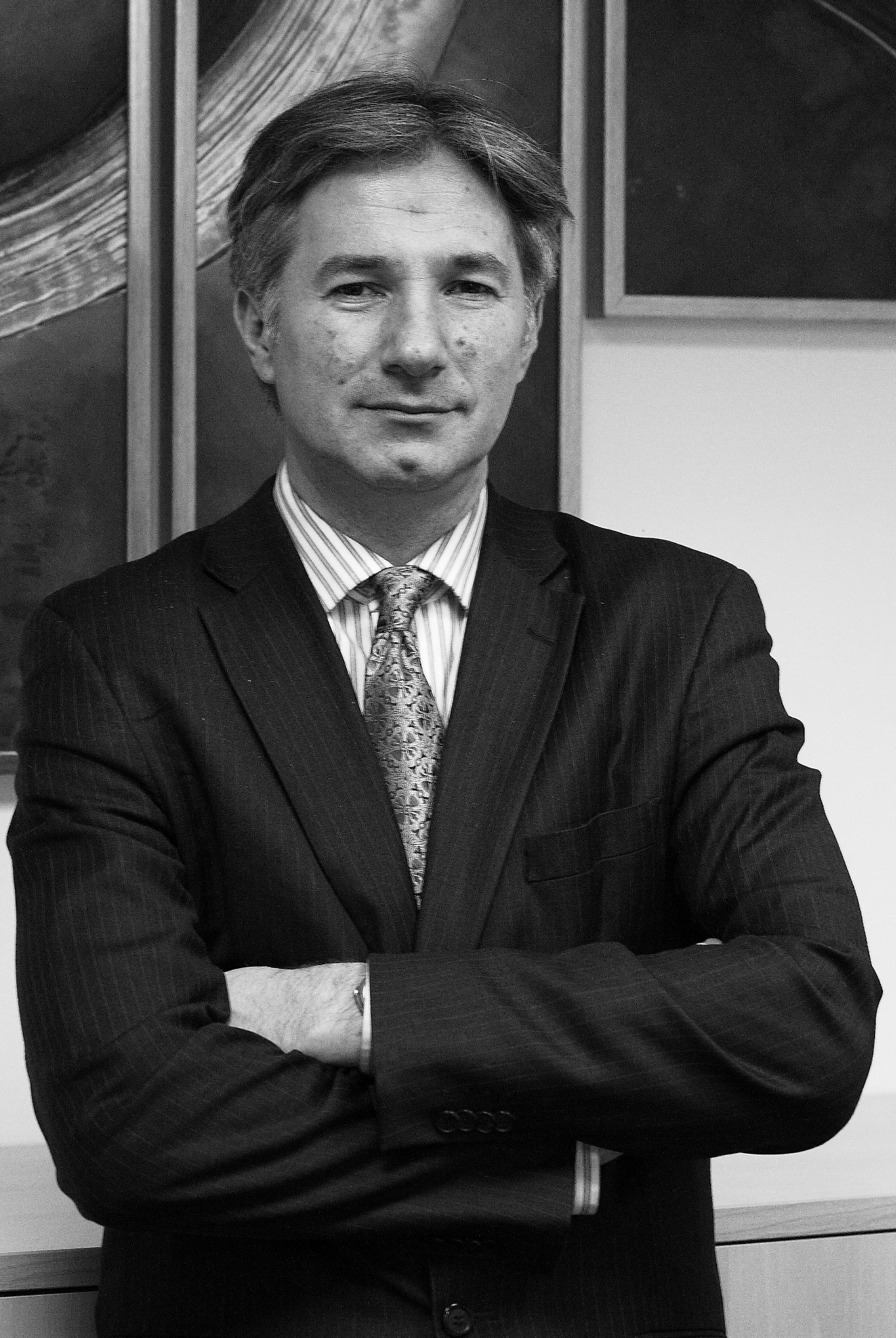Portret biznesowy mężczyzny w garniturze w odcieniach szarości, z założonymi rękami na tle ściany z obrazami.