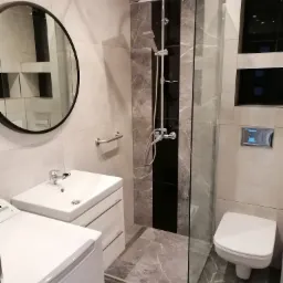 Nowoczesna łazienka z kabiną prysznicową typu walk-in, umywalką z szafką, okrągłym lustrem i pralką, wykończona płytkami imitującymi marmur z ciemnym pasem w kabinie.