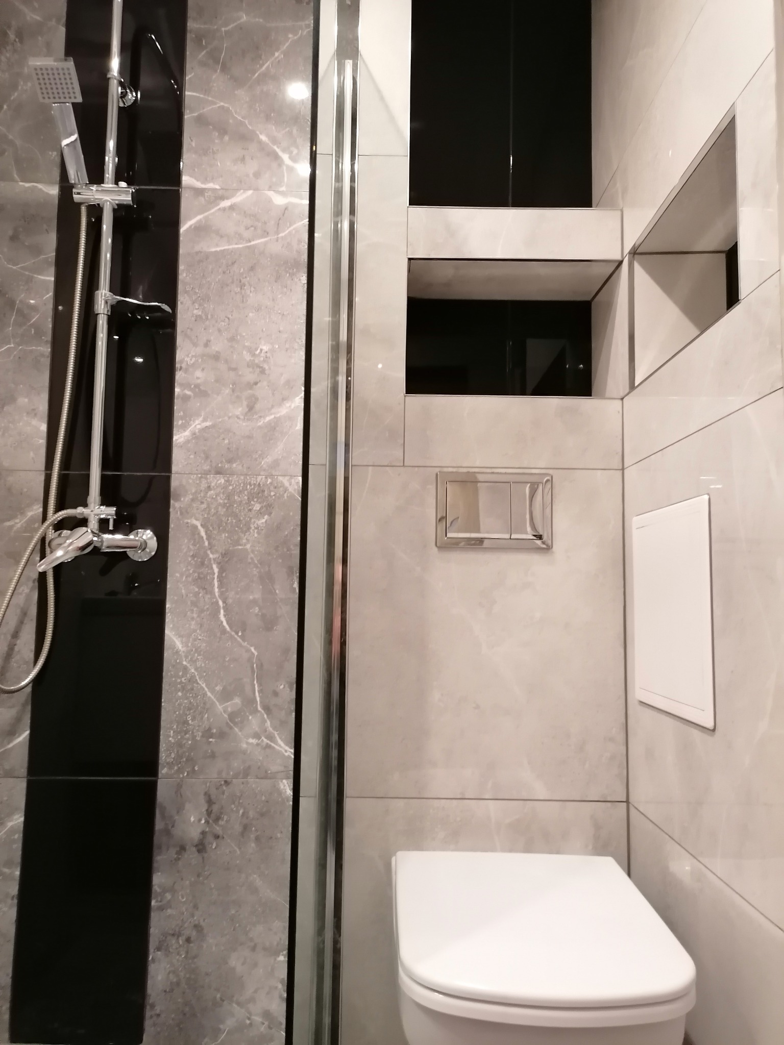 Nowoczesna łazienka z szarymi płytkami imitującymi marmur, kabiną prysznicową ze srebrnymi profilami, wiszącą toaletą i wnękami w ścianie.