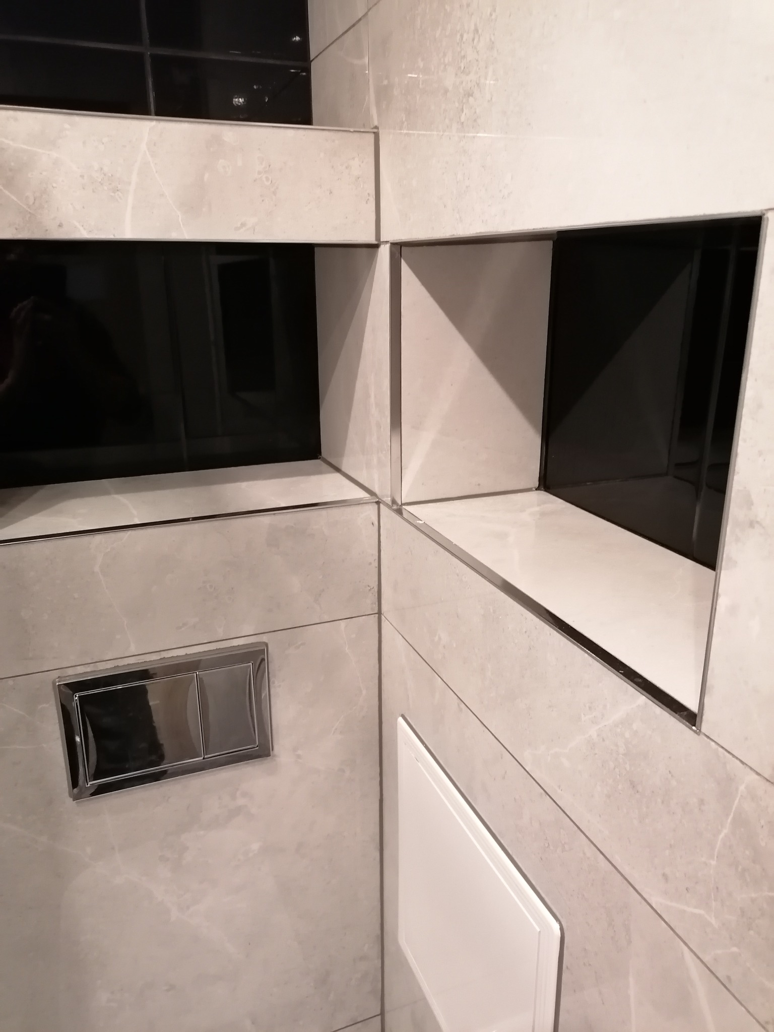 Wykończona ściana łazienki z szarych płytek imitujących marmur, z wnękami wyłożonymi czarnymi płytkami i chromowanym przyciskiem spłuczki.