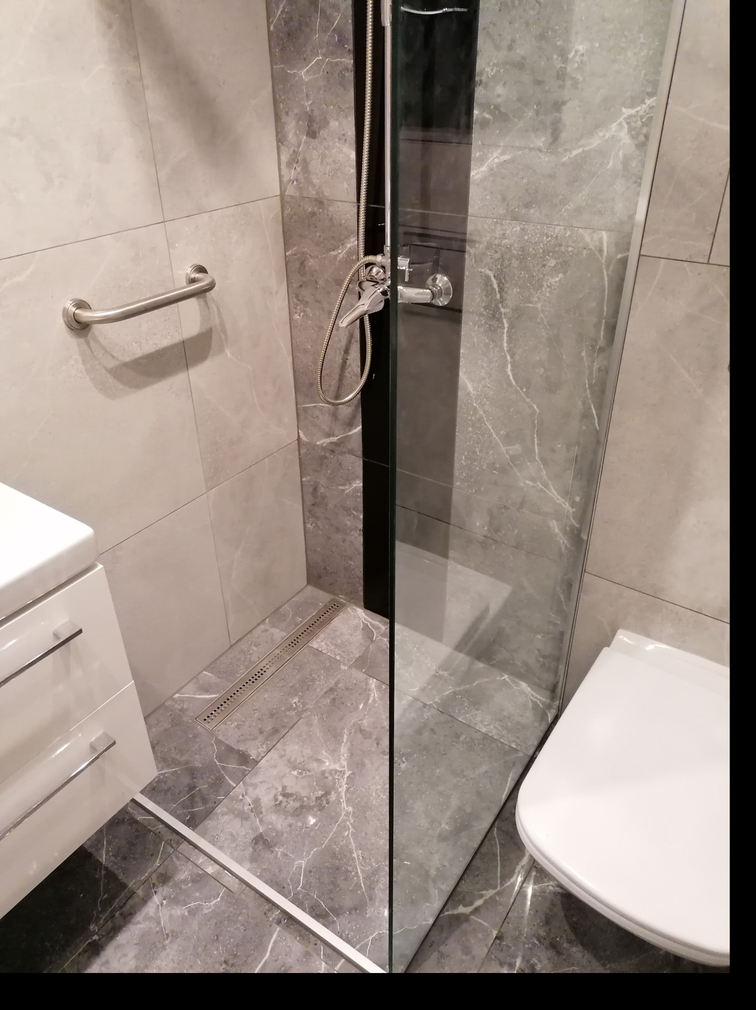 Nowoczesna łazienka z szarymi płytkami imitującymi marmur, kabina prysznicowa ze szklaną ścianką, odpływ liniowy, biała szafka z umywalką i chromowany uchwyt dla niepełnosprawnych.