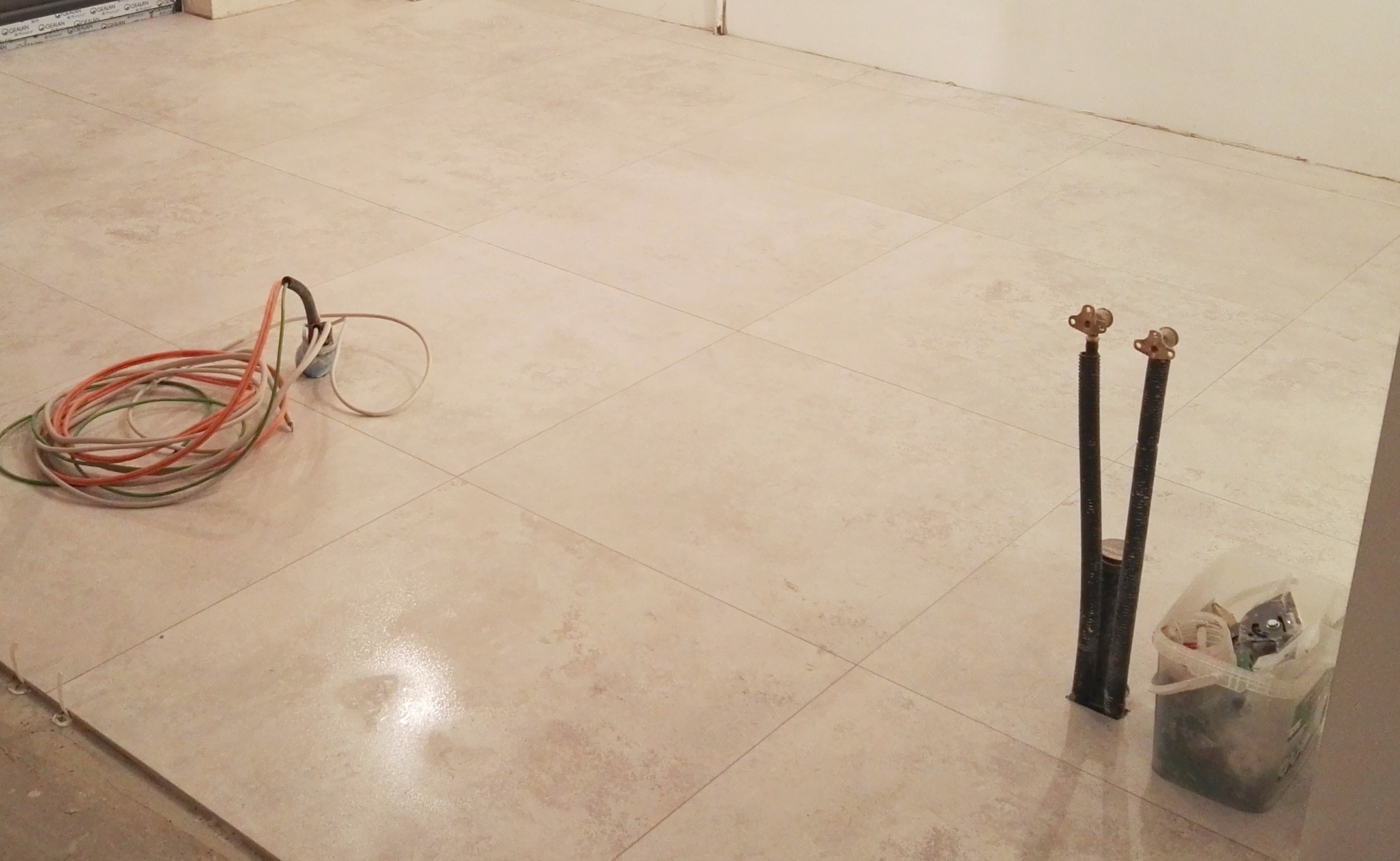 Ułożona, jasna, kwadratowa podłoga z płytek ceramicznych z widocznymi rurami instalacji wodnej i zwiniętym kablem elektrycznym oraz pojemnikiem z narzędziami.