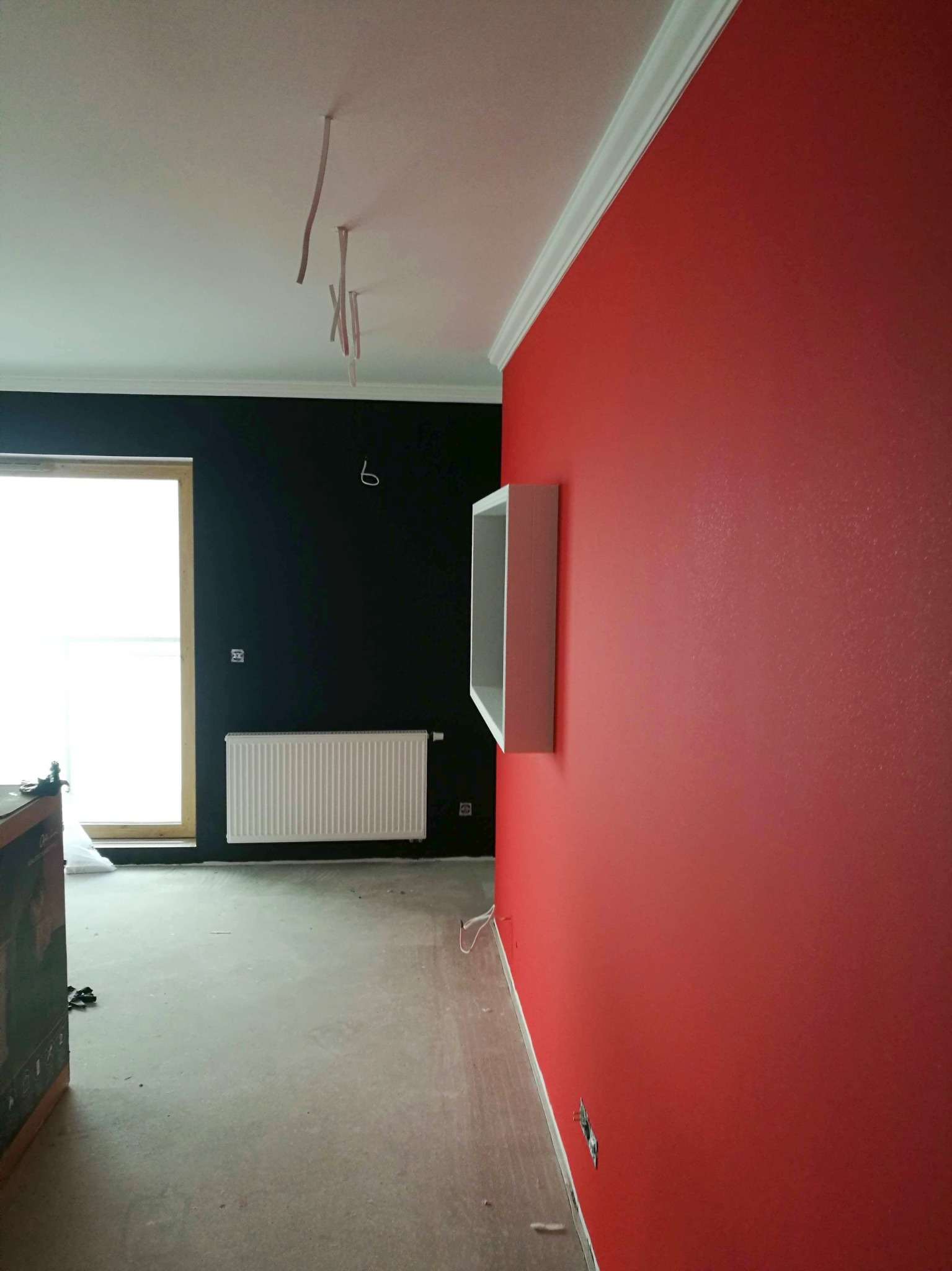 Świeżo pomalowane wnętrze z czerwoną i czarną ścianą, widoczny biały grzejnik, listwa sufitowa i wystające kable elektryczne, podłoga w trakcie wykańczania.