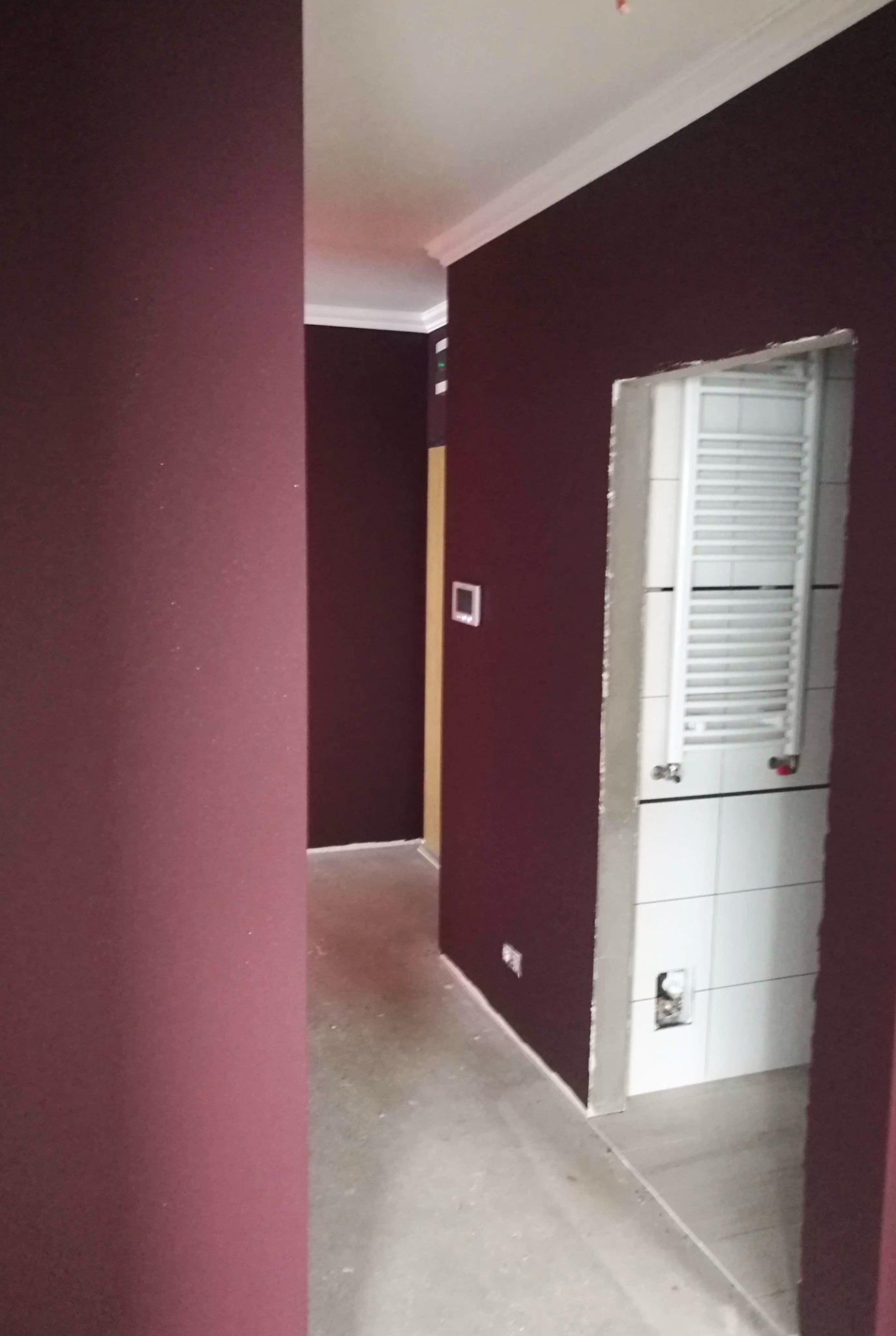 Korytarz z pomalowanymi na bordowo ścianami i wykończoną łazienką z białymi płytkami i grzejnikiem drabinkowym w trakcie remontu.