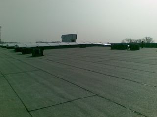 Płaski dach budynku przemysłowego pokryty papą termozgrzewalną, widoczne kominki wentylacyjne i elementy konstrukcyjne dachu. Perspektywa z lekkiego ukosa, w tle zamglone niebo.