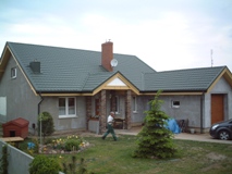 Dom jednorodzinny z zielonym dachem z dachówki, widoczne obróbki blacharskie, rynny w kolorze dachu oraz fragment ogrodu z tujami i budą dla psa. Na podjeździe zaparkowany samochód.