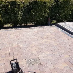 Ułożona kostka brukowa w odcieniach czerwieni i szarości, obrzeża z ciemnej kostki, widoczny właz kanalizacyjny oraz zwinięty przedłużacz na powierzchni, w tle żywopłot z tui.