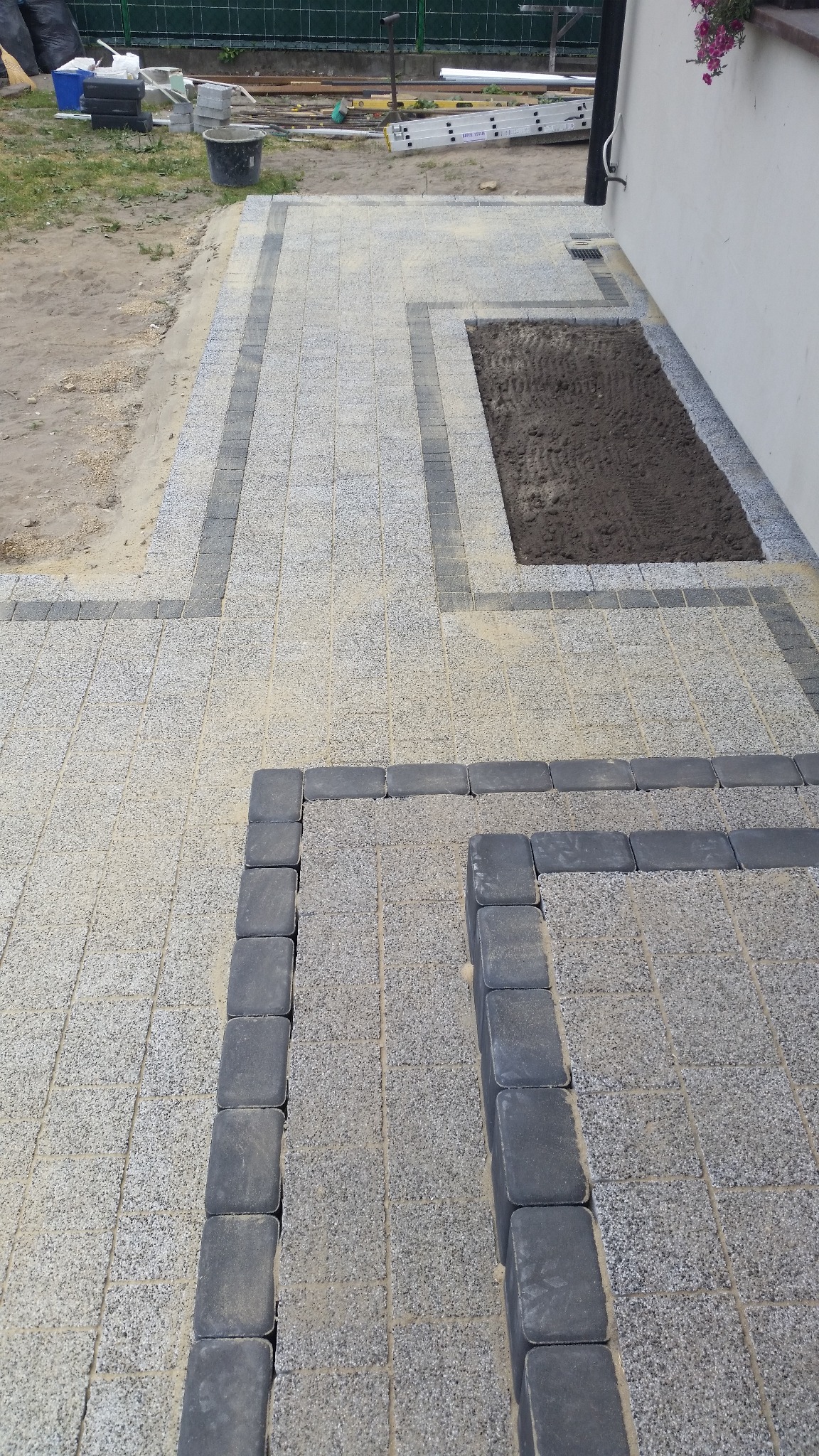 Ułożona szara kostka brukowa z ciemniejszym obramowaniem, fragment rabaty z ziemią, widoczny fragment budynku i drabina oparta o ścianę.