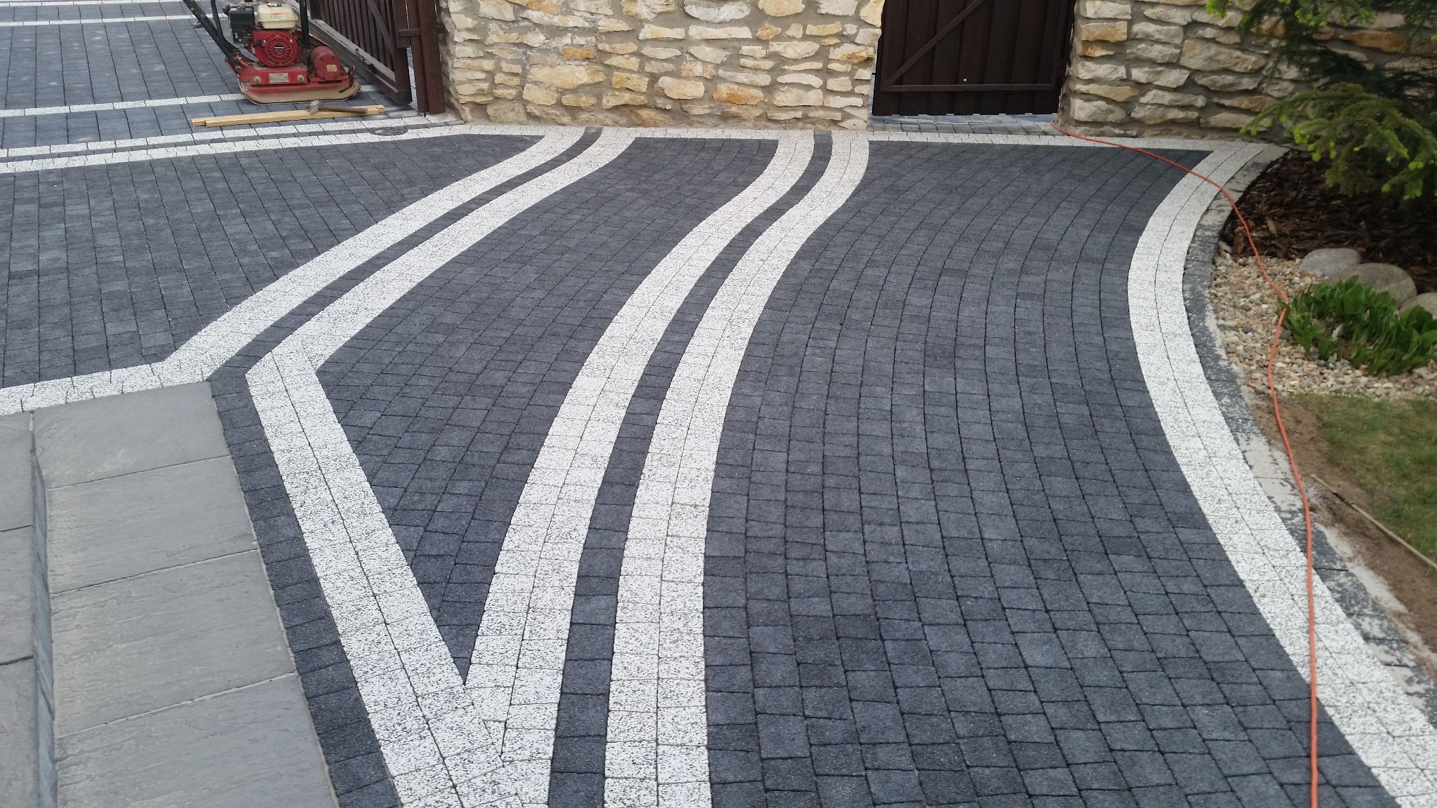 Ułożona kostka brukowa w kolorze grafitowym z białymi pasami, tworzącymi wzór na podjeździe, widoczna zagęszczarka w tle.
