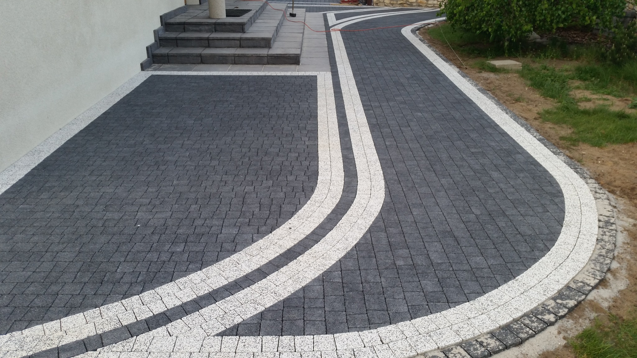 Ułożona, ciemnoszara kostka brukowa z białymi pasami dekoracyjnymi, prowadząca do schodów wejściowych, z widocznym fragmentem trawnika i elewacji budynku.