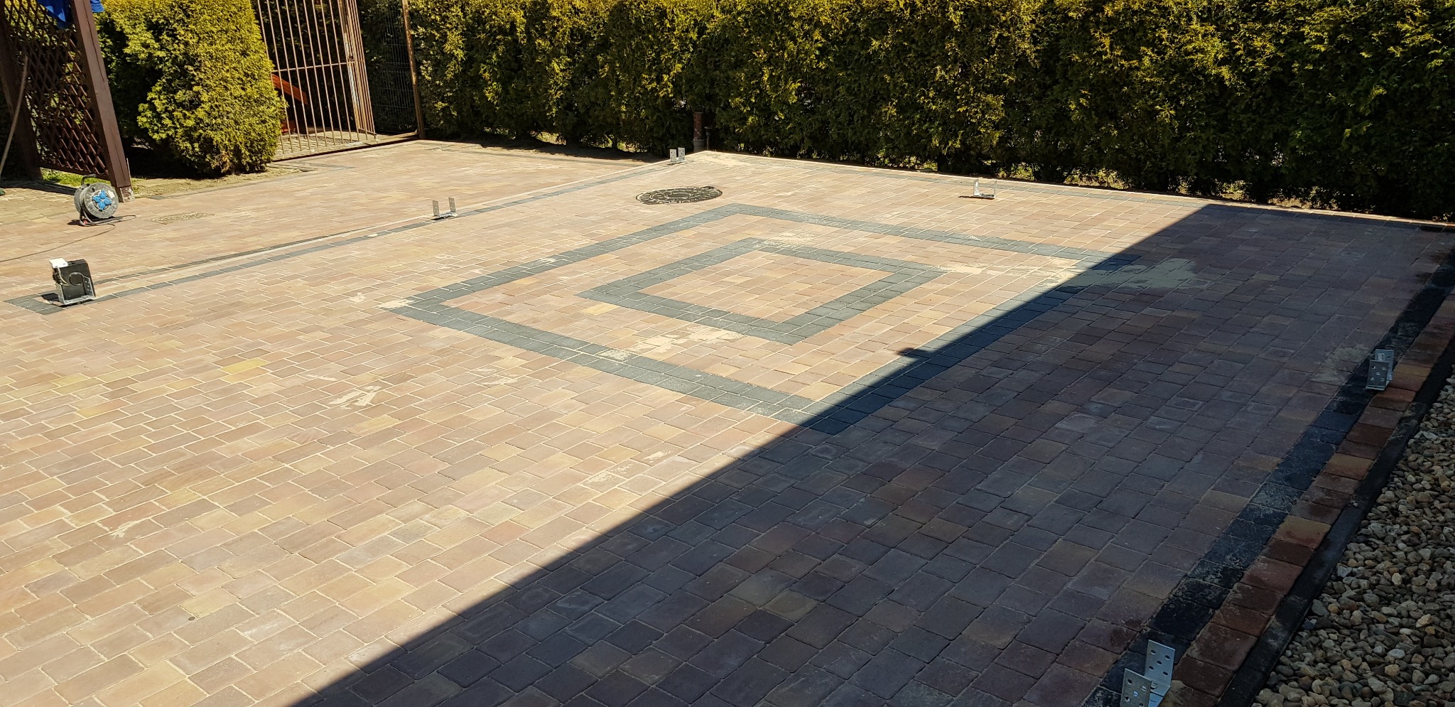 Ułożona kostka brukowa w Lędzinach z wzorem w kształcie kwadratu, otoczona zielonym żywopłotem, widoczne elementy oświetlenia i przyłącza elektryczne.
