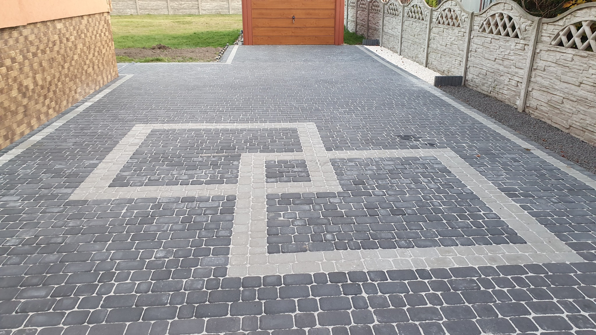 Ułożona kostka brukowa w kolorach szarości z jasnymi wzorami geometrycznymi przed budynkiem z brązową elewacją i drewnianym garażem, widoczne ogrodzenie z prefabrykatów betonowych.