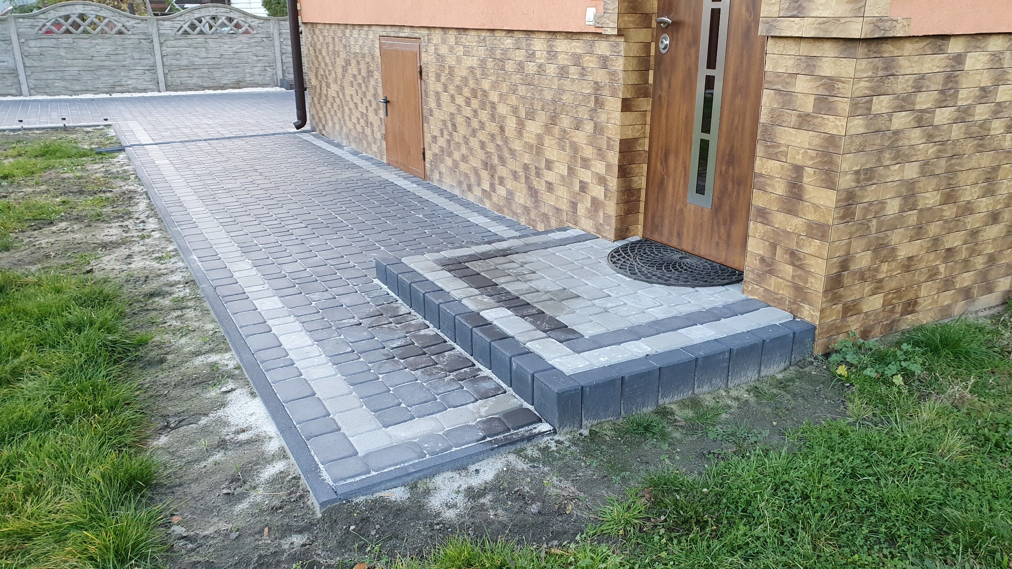 Ułożona szara kostka brukowa tworząca chodnik i schody wejściowe do domu z brązową elewacją, widoczny betonowy płot w tle.