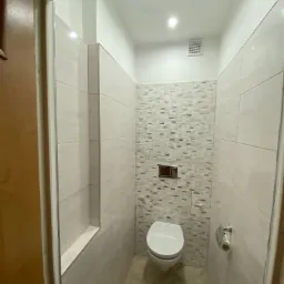 Wąska łazienka po remoncie z mozaikową ścianą, podwieszaną toaletą i beżowymi płytkami podłogowymi, widziana zza framugi drzwi.