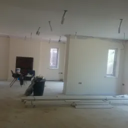 Wnętrze w trakcie malowania ścian na jasny kolor, widoczne listwy przypodłogowe, przygotowane do montażu, oraz instalacje elektryczne pod sufitem