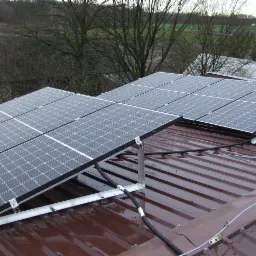 Panel fotowoltaiczny zamontowany na brązowym, metalowym dachu z widocznymi kroplami deszczu, z szarą konstrukcją wsporczą i czarnymi kablami w peszlach, w tle drzewa bez liści i zabudowania.