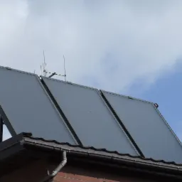 Trzy kolektory słoneczne zamontowane na dachu budynku z brązową blachodachówką i anteną telewizyjną na tle błękitnego nieba z chmurami.