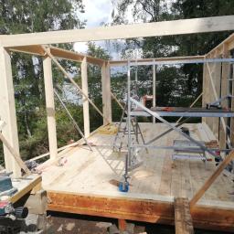 Paweł Mołczan - Wznoszenie szkieletu drewnianego budynku, widoczne deski, rusztowanie, narzędzia do cięcia drewna oraz elementy izolacyjne wewnątrz konstrukcji, tło stanowią drzewa i częściowo widoczne jezioro.