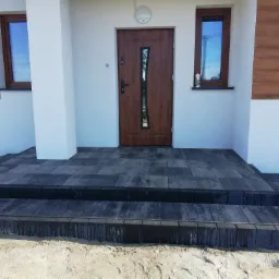 Nowoczesne wejście do domu z ciemnoszarą kostką granitową ułożoną na schodach i podeście, biała elewacja z drewnopodobnymi panelami, brązowe okna i drzwi z pionowym przeszkleniem.