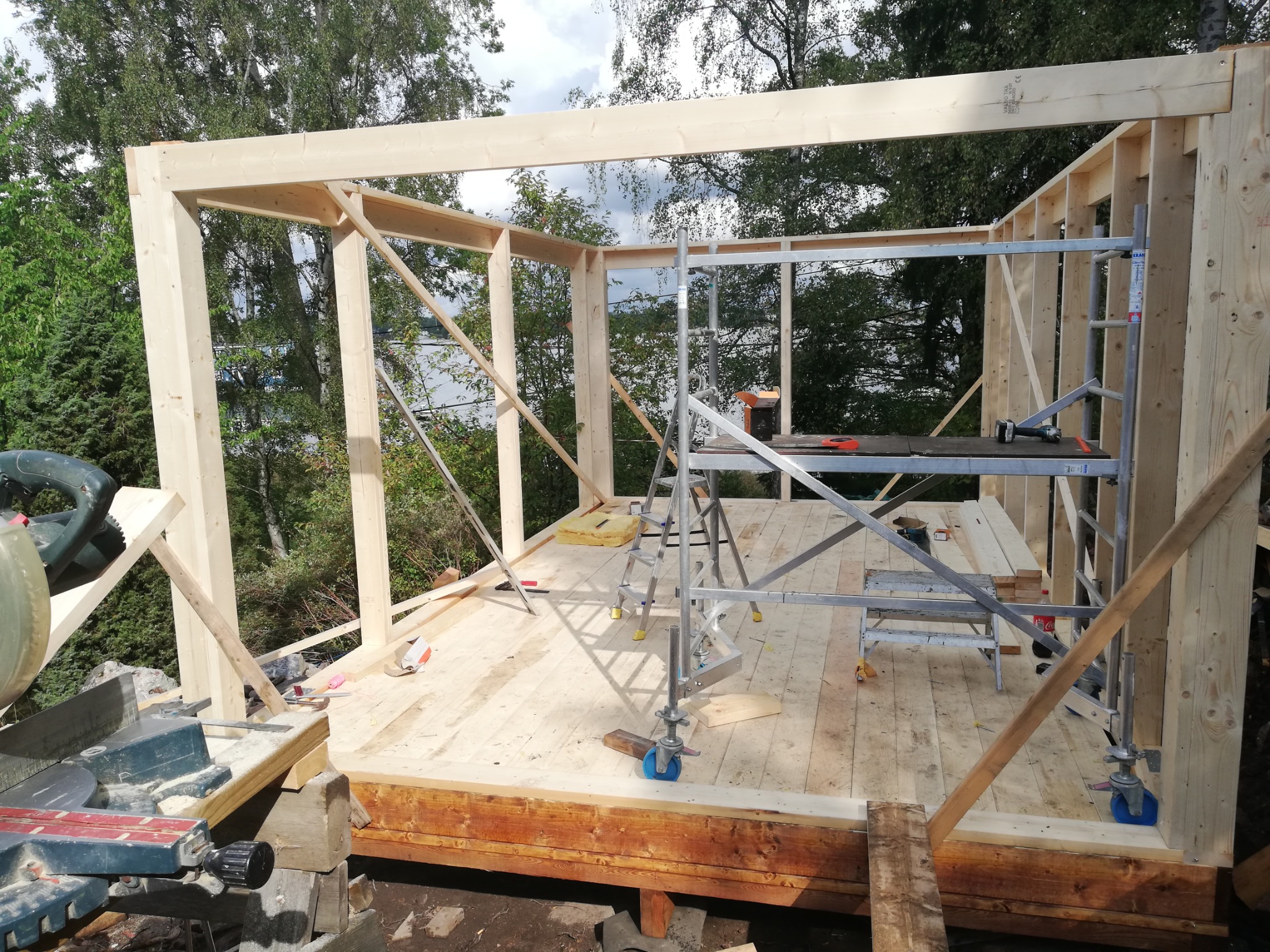 Wznoszenie szkieletu drewnianego budynku, widoczne deski, rusztowanie, narzędzia do cięcia drewna oraz elementy izolacyjne wewnątrz konstrukcji, tło stanowią drzewa i częściowo widoczne jezioro.