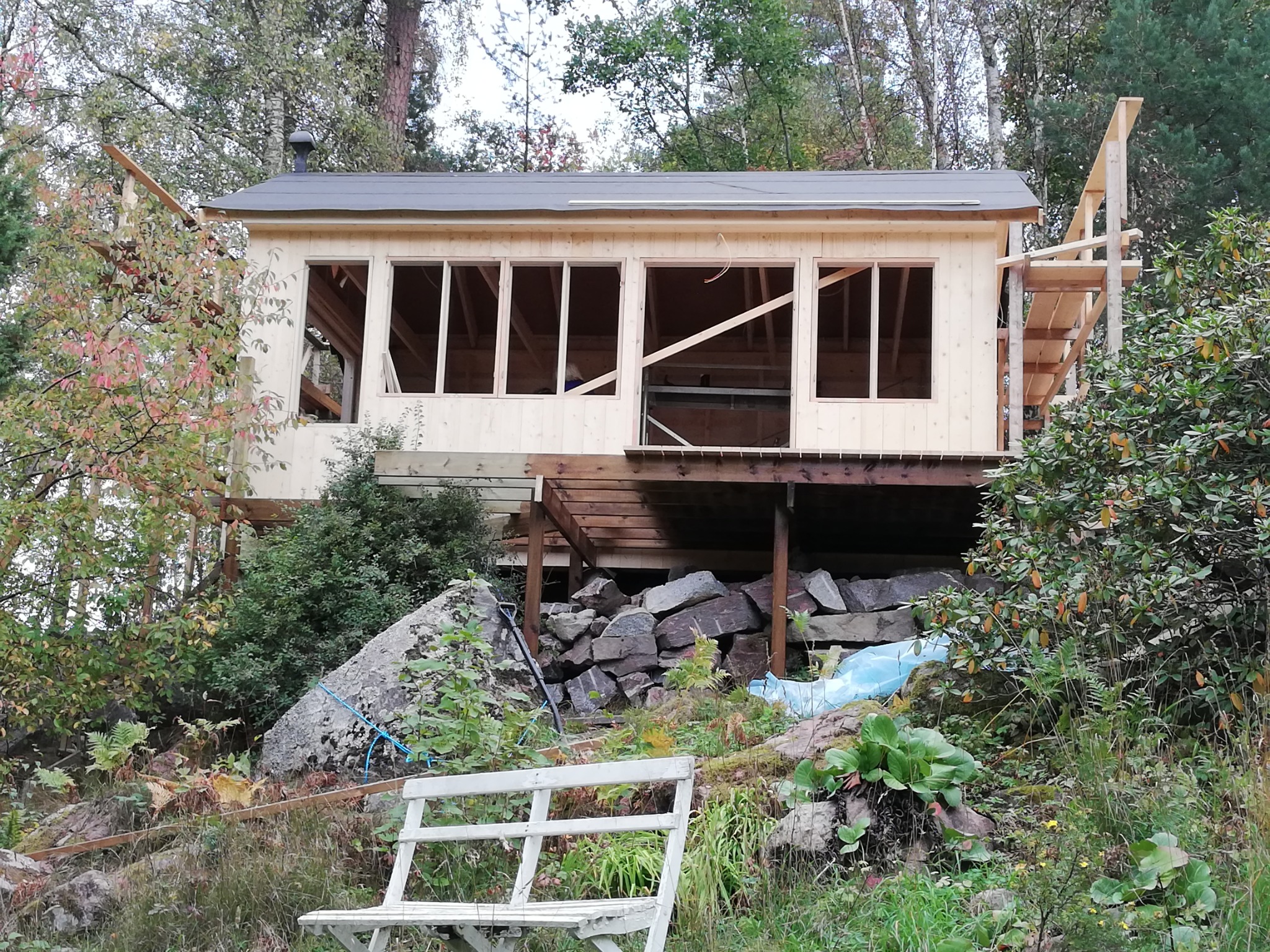 Wznoszony drewniany budynek z płaskim dachem na skarpie, widoczne niezabudowane otwory okienne, drewniany pomost i rusztowanie, w tle zieleń i biała ławeczka.