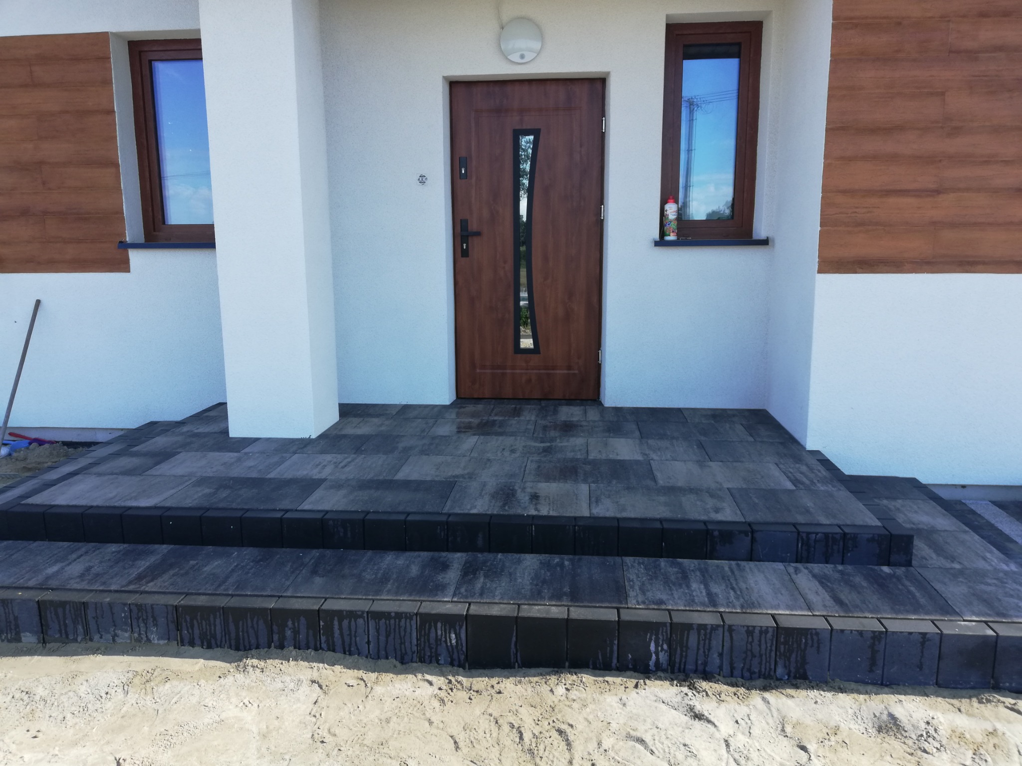 Nowoczesne wejście do domu z ciemnoszarą kostką granitową ułożoną na schodach i podeście, biała elewacja z drewnopodobnymi panelami, brązowe okna i drzwi z pionowym przeszkleniem.