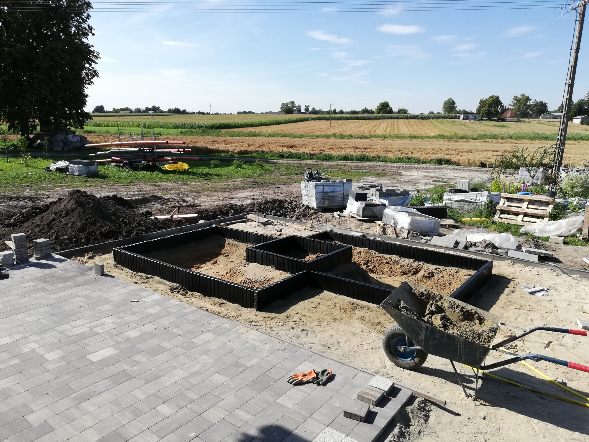 Budowa tarasu z kostki brukowej w trakcie realizacji, widoczny zarys rabat kwiatowych, taczka z piaskiem, ułożona część nawierzchni i stosy kostki brukowej na tle pola.