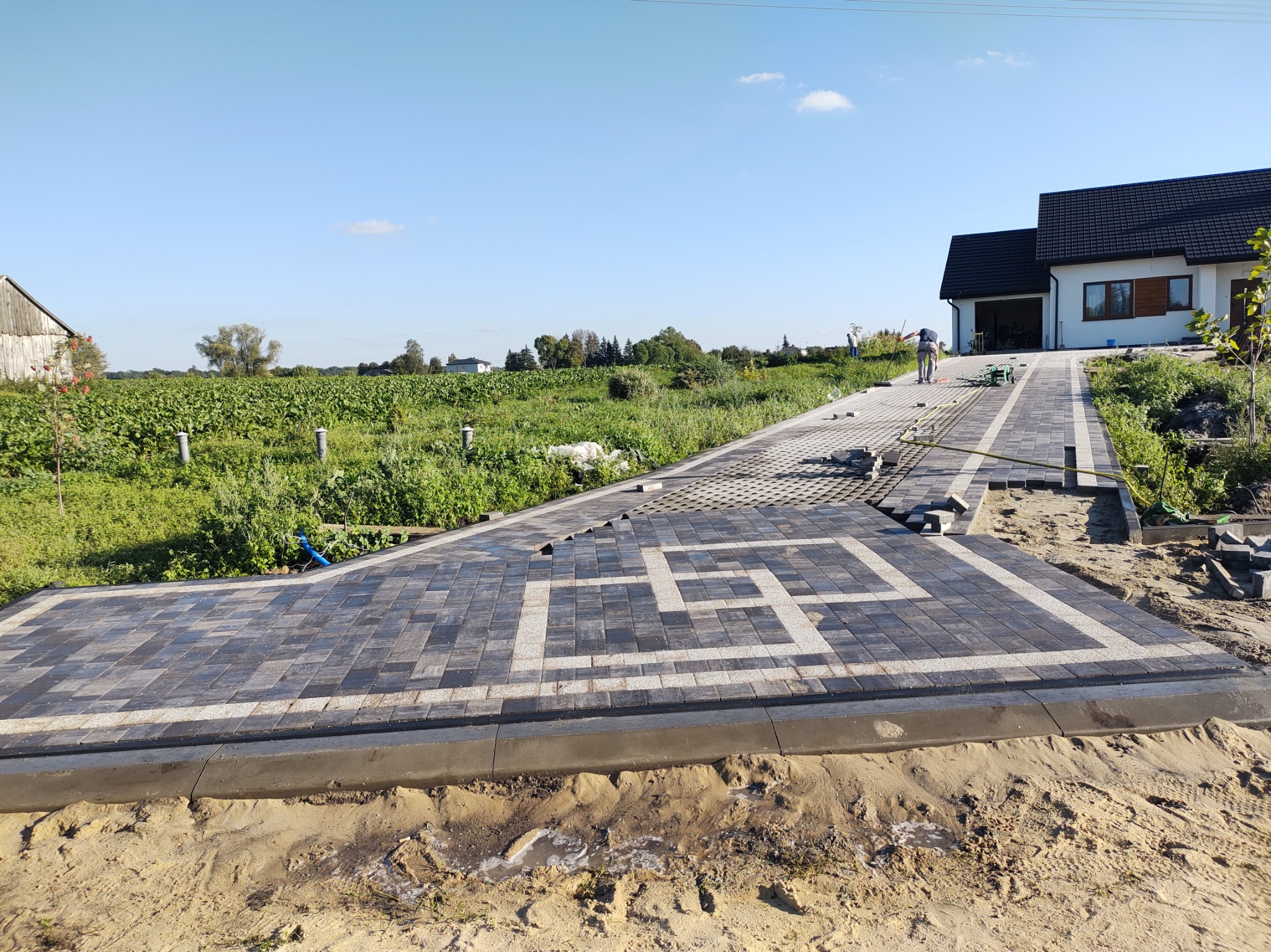 Układanie wzoru z ciemnoszarej kostki brukowej z białymi liniami na podjeździe do domu, widoczne niezakończone fragmenty i pracownik w oddali.