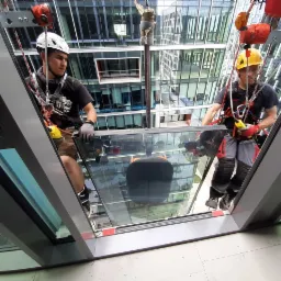 Dwóch pracowników na wysokościach, w kaskach i uprzężach, montuje dużą szklaną taflę na elewacji biurowca, widoczny system lin i zabezpieczeń.