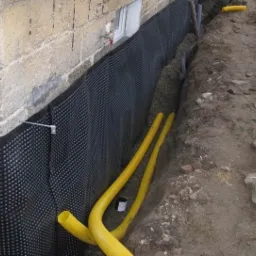 Fundament budynku zabezpieczony czarną folią kubełkową, widoczne żółte rury drenażowe w wykopie, jasna elewacja budynku z małym oknem.