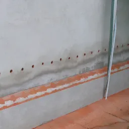 Świeżo otynkowana ściana fundamentowa z rzędem nawierconych otworów wentylacyjnych i zabezpieczoną warstwą hydroizolacji na styku z posadzką.