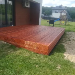 Nowoczesny taras z drewna w odcieniach brązu, przylegający do domu z klimatyzatorem na elewacji, z widokiem na ogród z grillem i placem zabaw w tle.