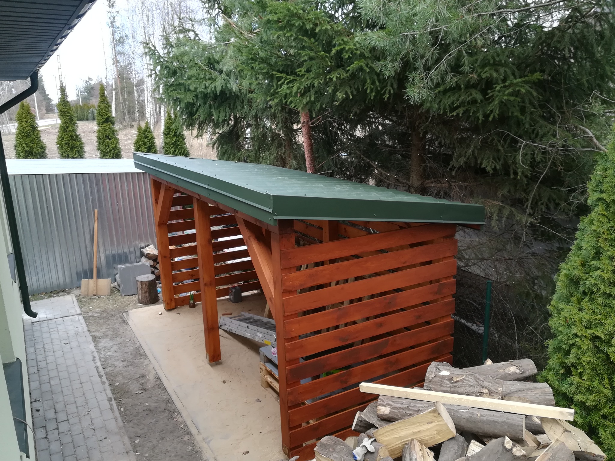 Drewniana konstrukcja z zielonym dachem, prawdopodobnie wiata na drewno, z ułożonymi polanami, na tle drzew i krzewów.