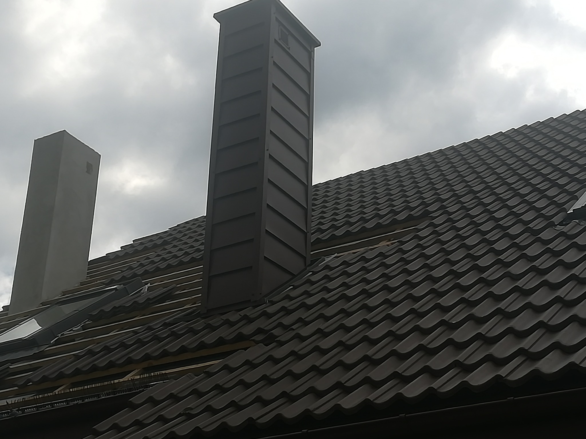 Montaż brązowej blachodachówki na dachu z drewnianą konstrukcją i dwoma kominami w pochmurny dzień.