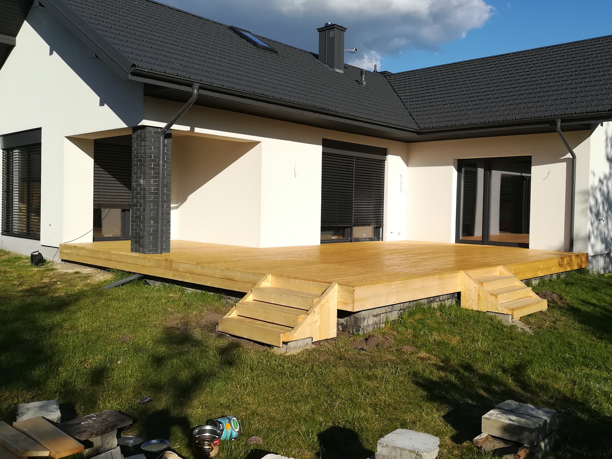 Nowoczesny dom z jasną elewacją i drewnianym tarasem z małymi schodkami, widoczne narzędzia i puszki z farbą na trawie, częściowo zacienione ujęcie.