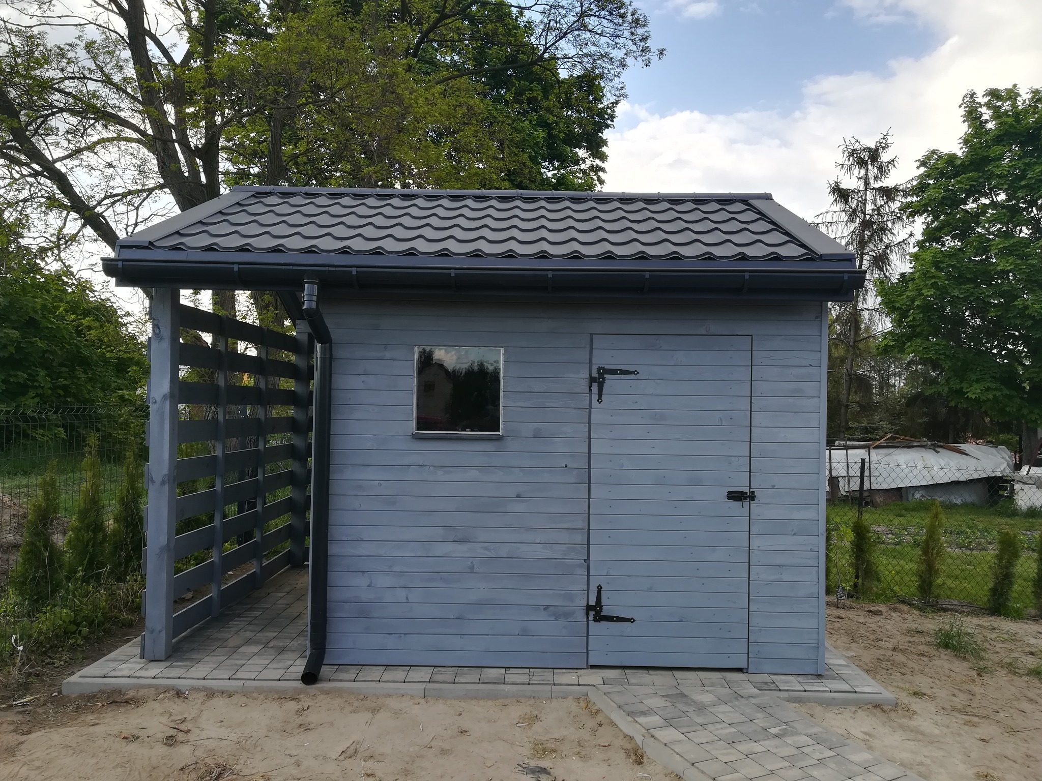 Szara drewniana budowla ogrodowa z metalowym dachem i małym oknem, widoczna z zewnątrz, z zadaszonym wejściem i brukowaną ścieżką.