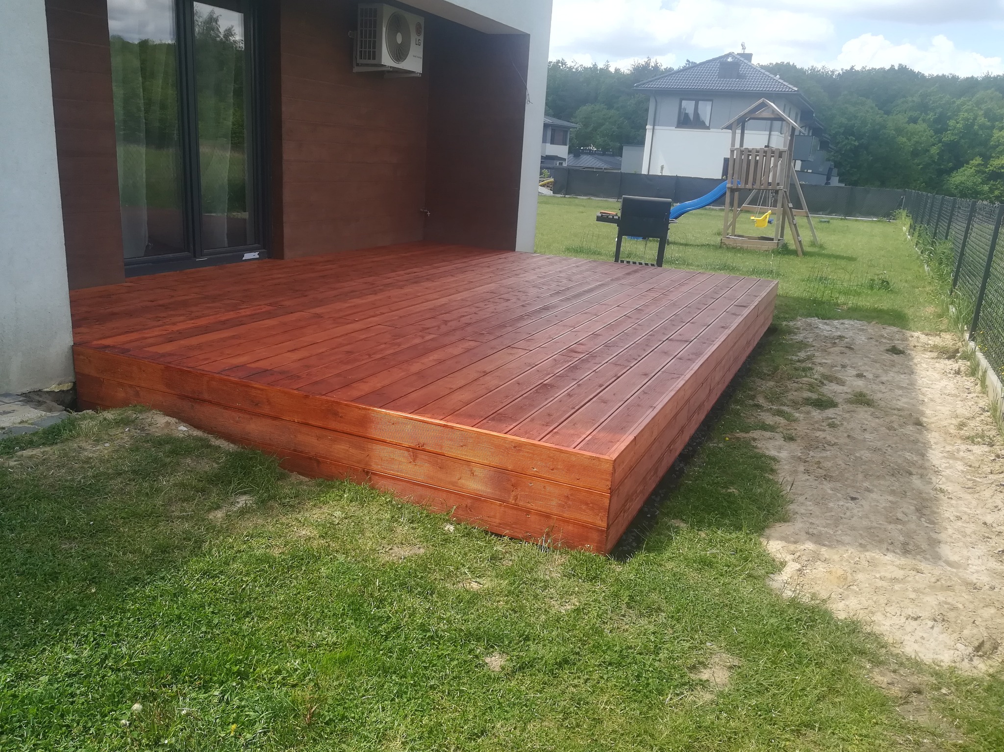 Nowoczesny taras z drewna w odcieniach brązu, przylegający do domu z klimatyzatorem na elewacji, z widokiem na ogród z grillem i placem zabaw w tle.