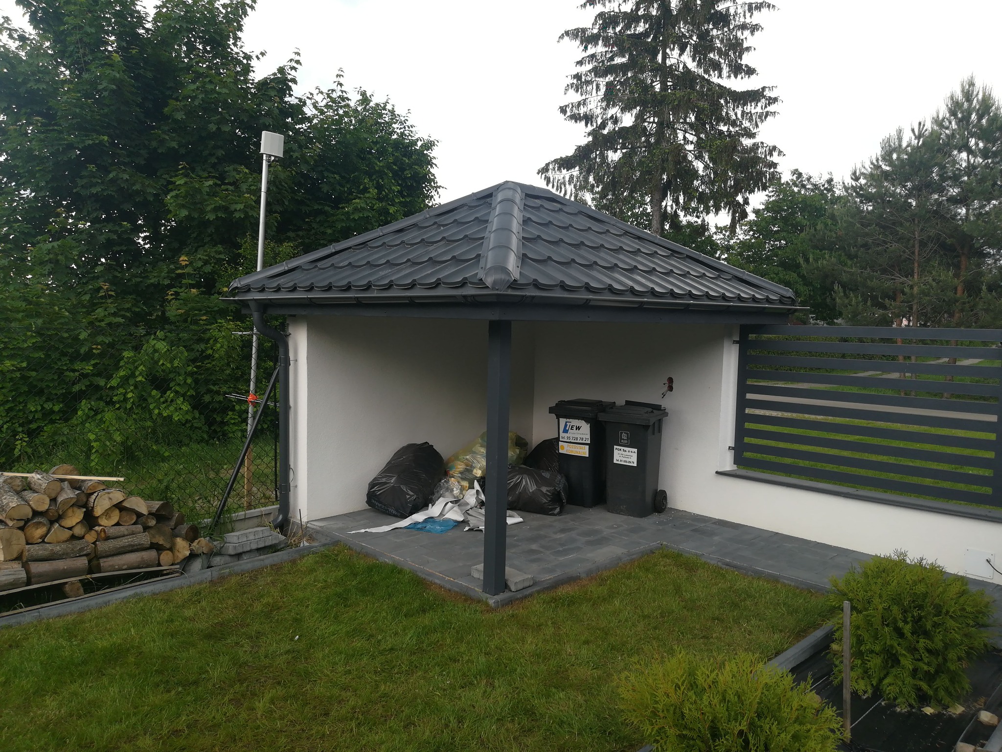 Zadaszona wiata śmietnikowa z szarą dachówką, obok składowane drewno opałowe, widoczne worki ze śmieciami i pojemniki na odpady na kostce brukowej.