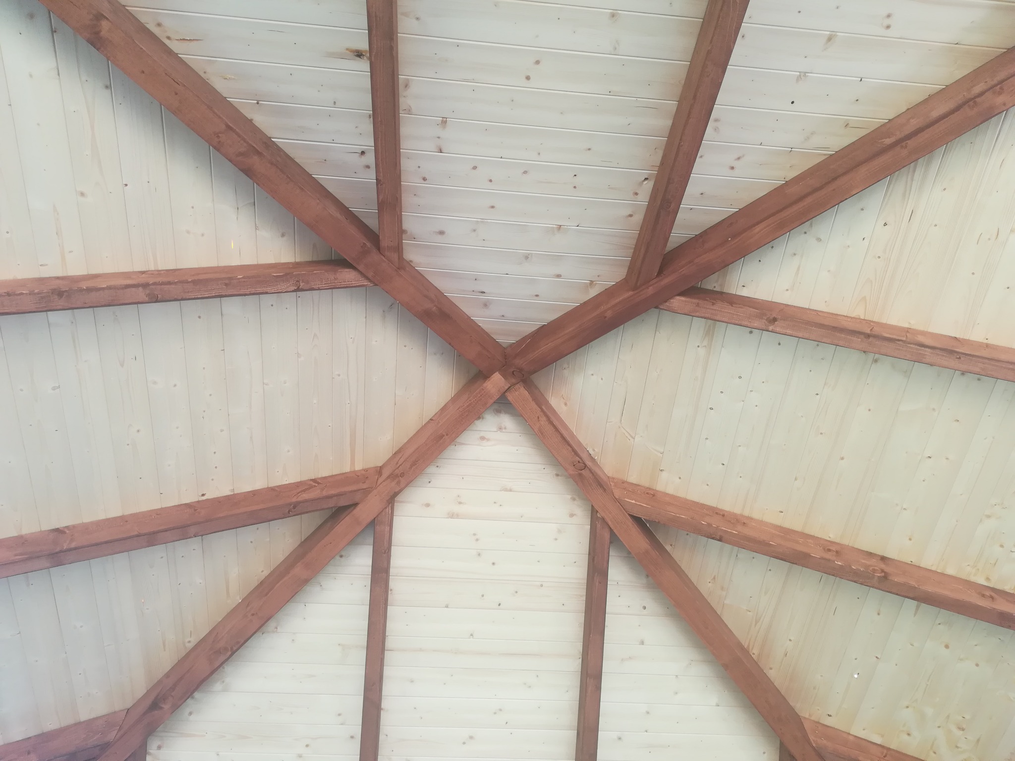 Drewniana konstrukcja dachu altany z brązowymi belkami na tle jasnego, drewnianego poszycia, widok z dołu.
