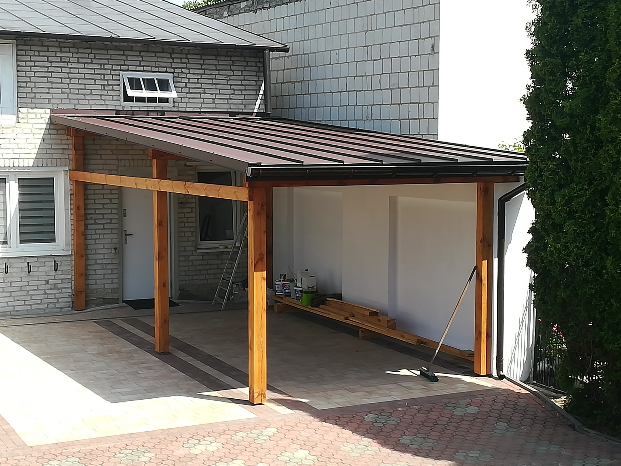 Zadaszenie tarasu z drewnianą konstrukcją i metalowym dachem, widoczne puszki z farbą i drabina oparta o ścianę budynku w tle.