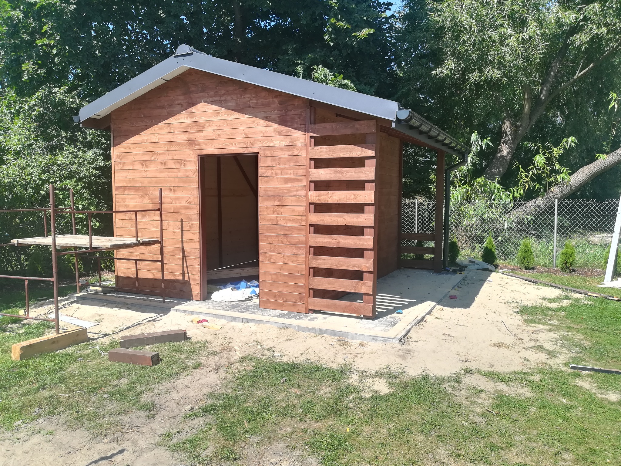 Drewniana konstrukcja z szarym dachem w trakcie budowy, widoczne rusztowanie, niezabezpieczone wejście i elementy konstrukcyjne z listew, otoczenie w stanie surowym.